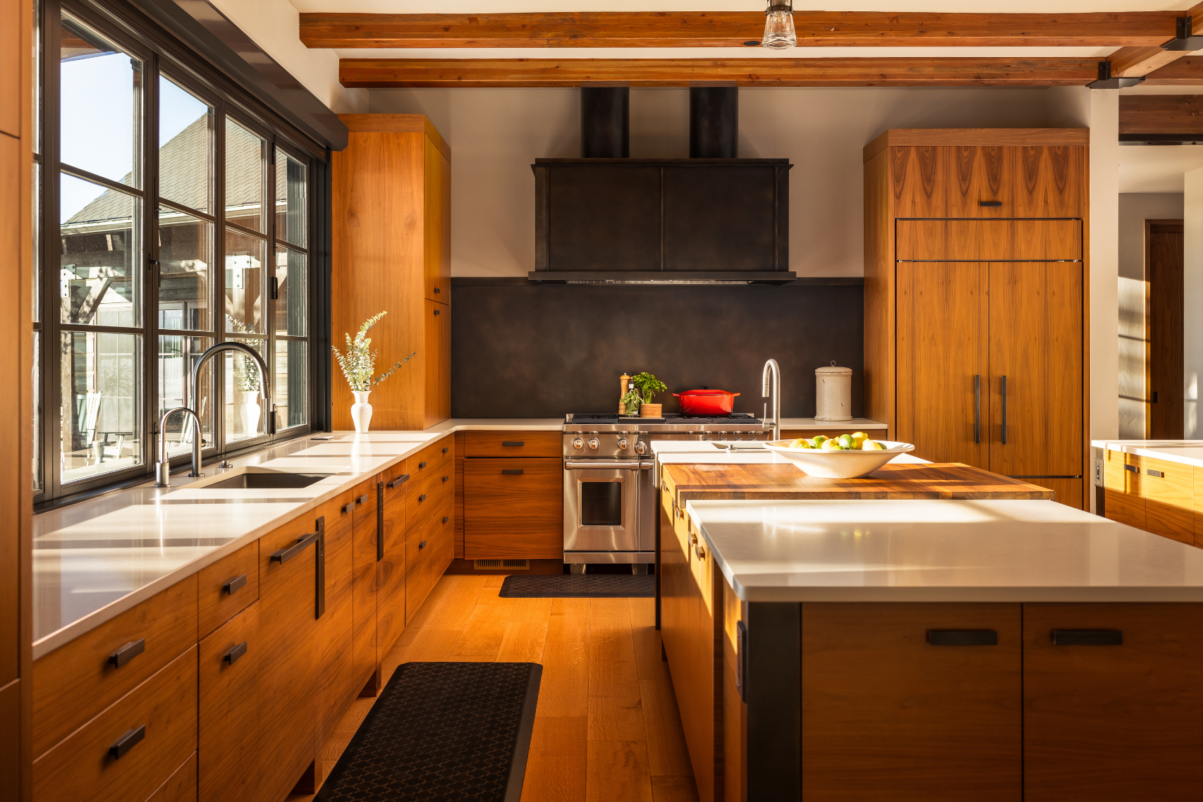 The sunny modern kitchen features 6’s wood cabinets, white countertops, a large island with a bowl of apples, stainless steel stove, black range hood, and wide windows that fill the space with natural light.