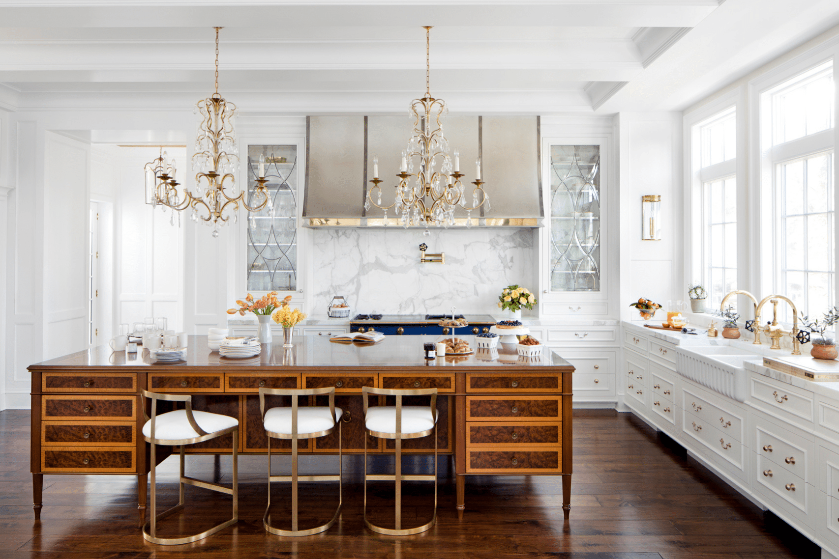 A luxurious kitchen features Product Name: 9—a large wooden island, three white stools with gold accents, marble backsplash, glass-front cabinets, two elegant chandeliers, a farmhouse sink, and expansive windows for natural light.