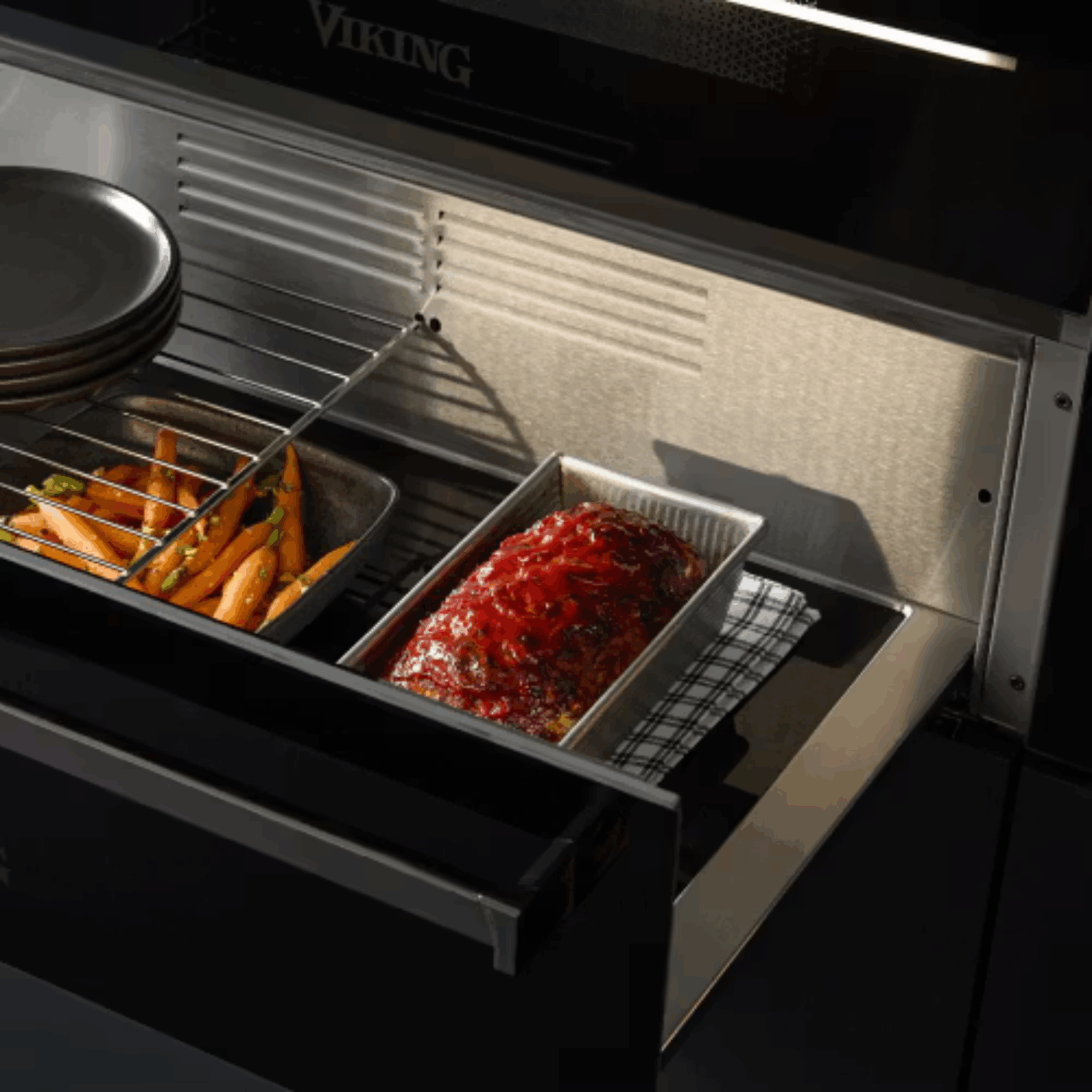 A Food warmer holds a tray of roasted carrots, a meatloaf topped with tomato sauce in a pan, and stacked plates on a rack inside its stainless steel compartment.