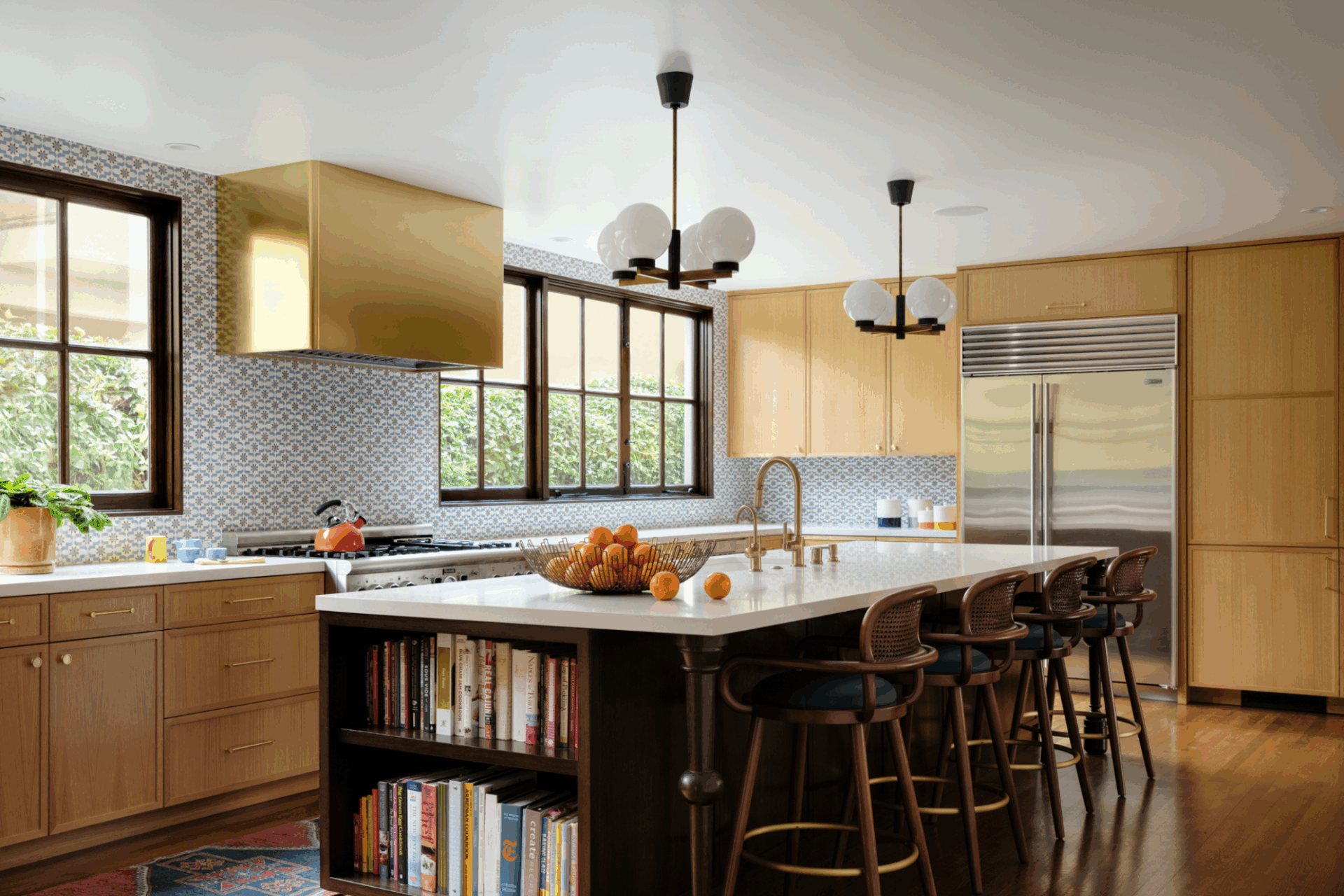 Modern kitchen featuring the L Shape with light wood cabinets, a long island with marble countertop, four barstools, open shelving for books, stainless steel appliances, gold vent hood, large windows, and decorative pendant lights.