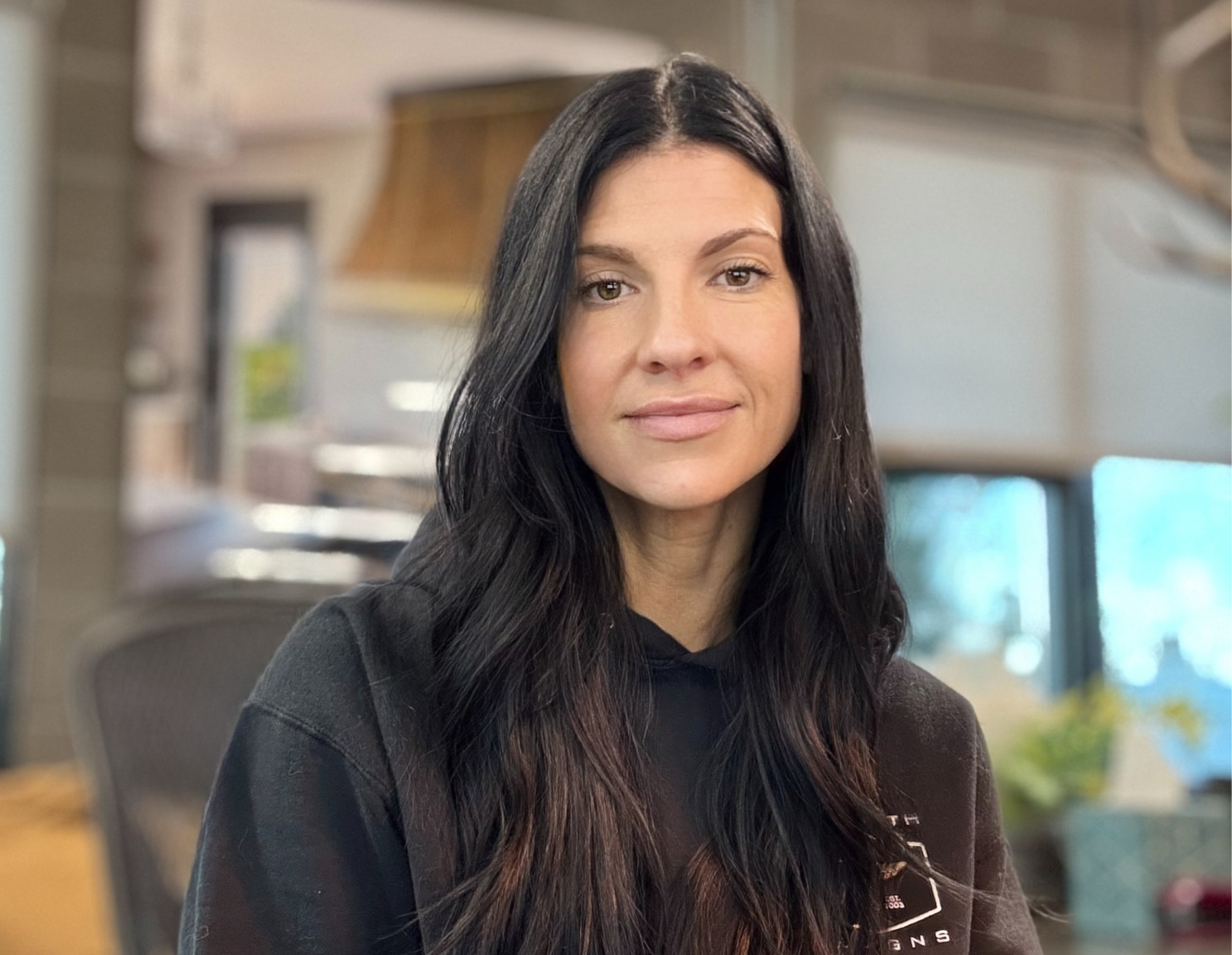 A woman with long brown hair, wearing a black top and necklace, stands smiling behind a counter. The Ella Bio-3 laptop and an office phone can be seen in the background.